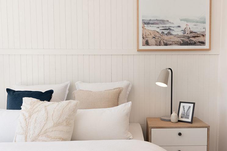 A calm coastal bedroom with layered cushions and a framed seaside print